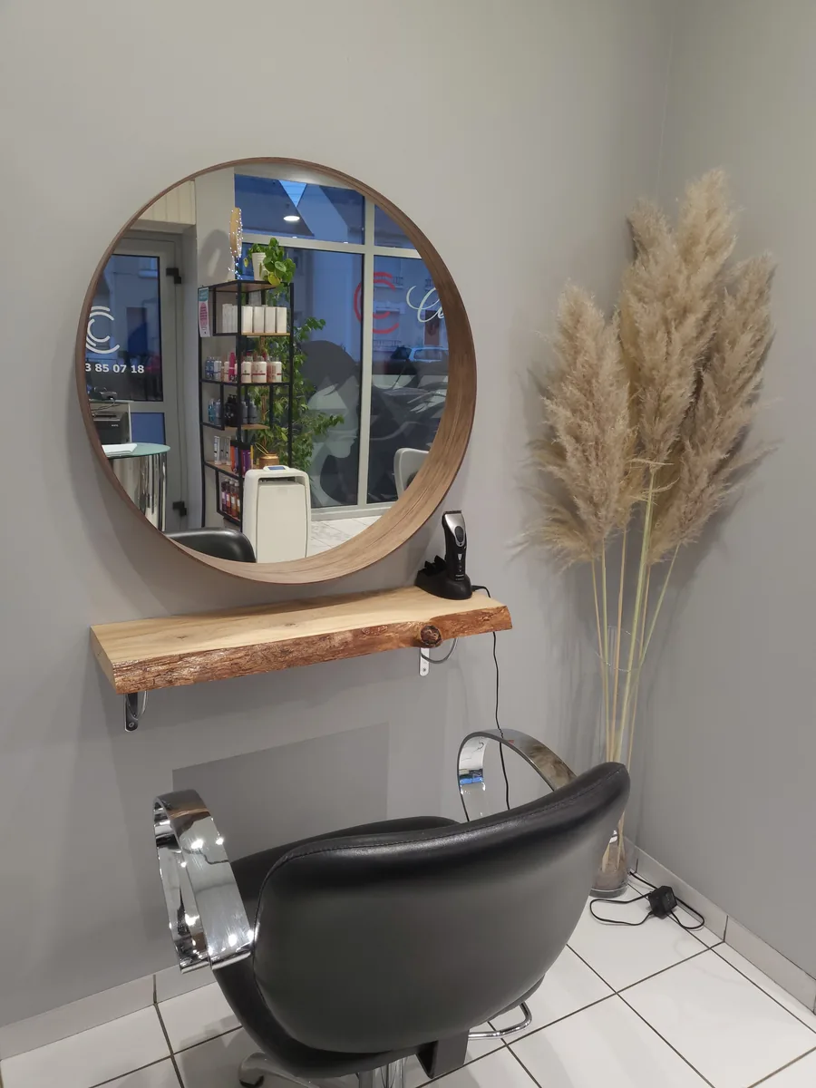 Un poste du salon : le miroir rond cerclé de bois, la tablette en bois brut, le fauteuil noir et les pampas